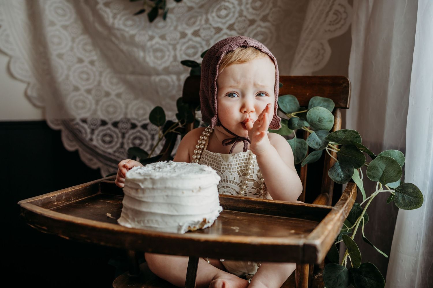 A small child in a high chair enjoying a white birthday cake with green foliage decor.