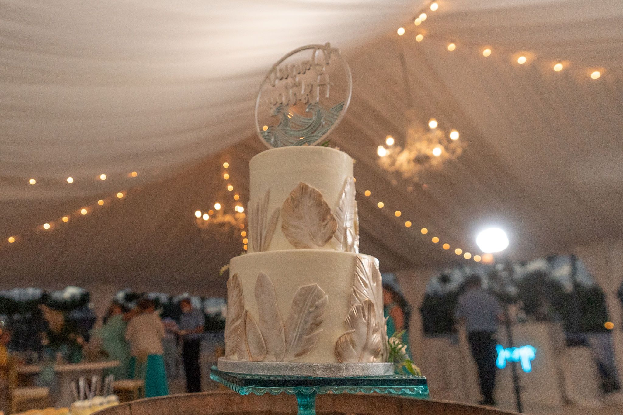Three-tiered white wedding cake with teal accents and decorative topper under string lights.