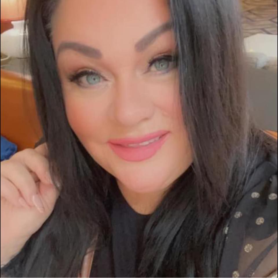 A woman with long black hair and blue eyes poses for a glamorous selfie wearing a black polka dot top.