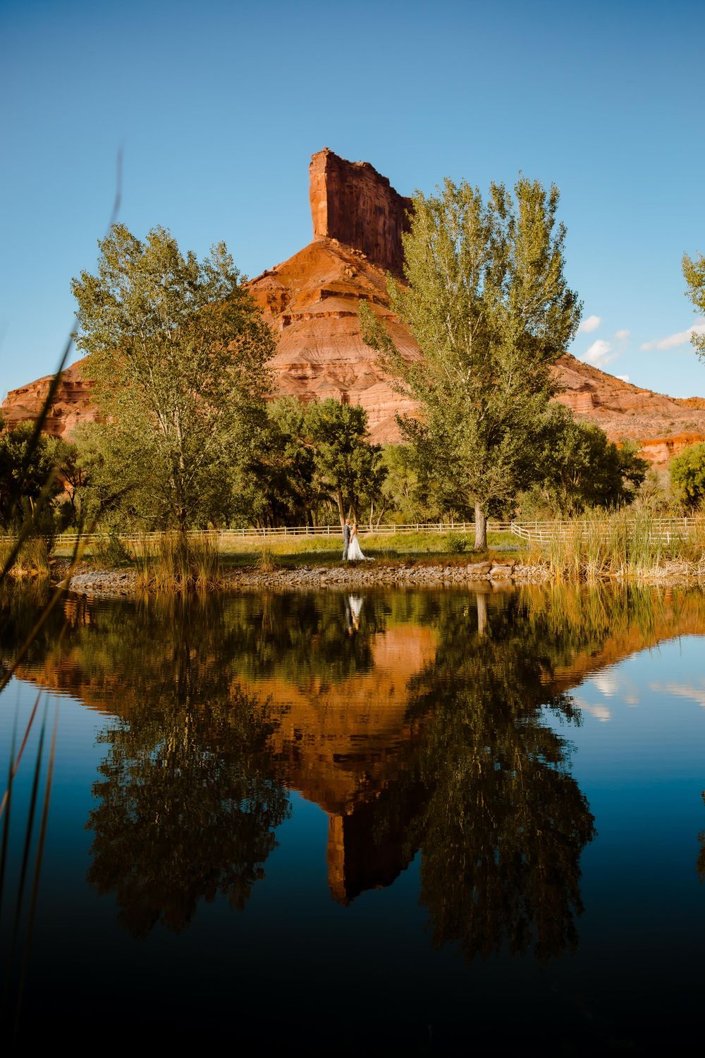 a bride and groom are reflected in a Colorado mountain lake with a chimney red rock and bright blue sky behind them