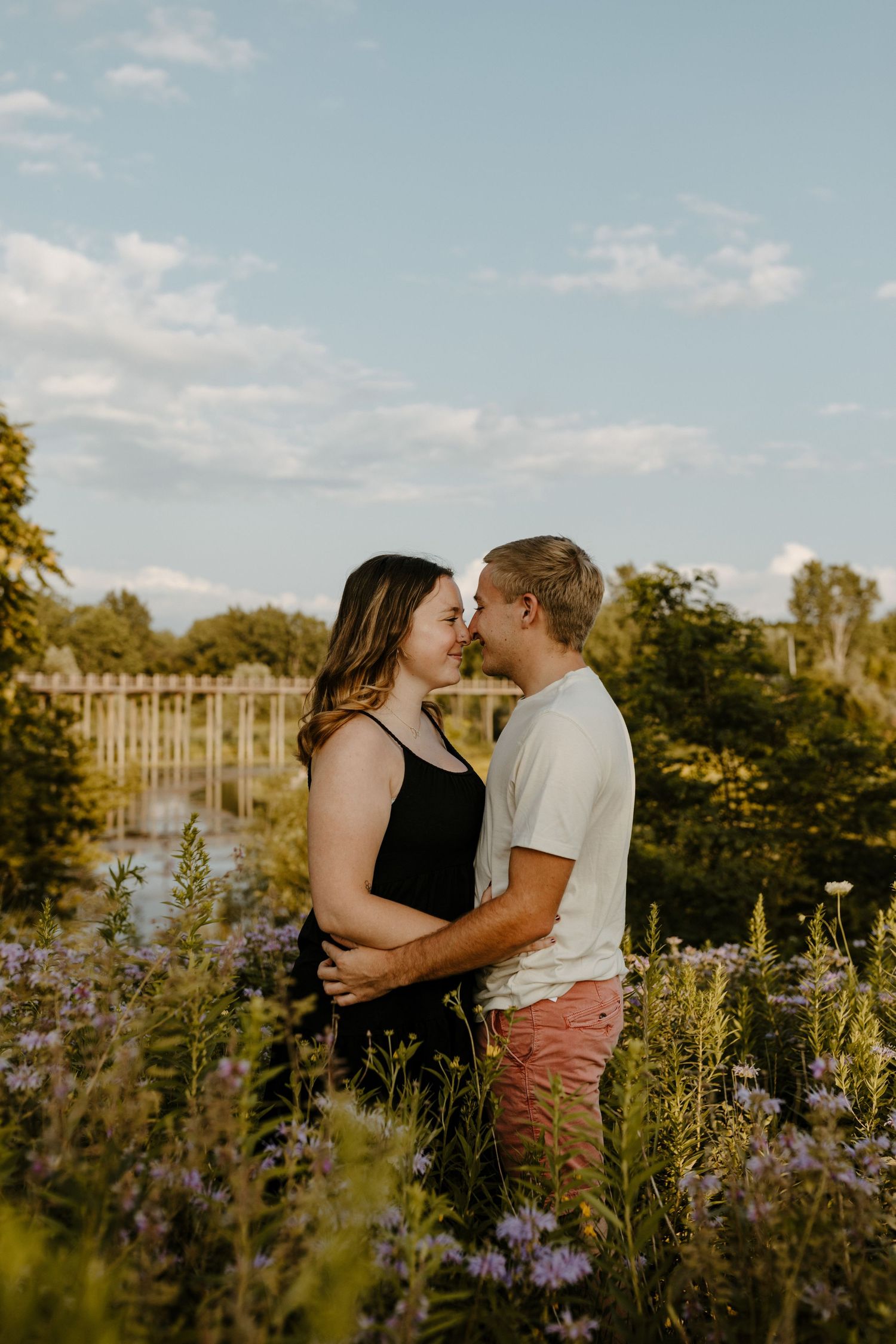 A couple embracing in a field of flowers, with a fence and trees in the background. Fort Worth Texas engagement photographer.