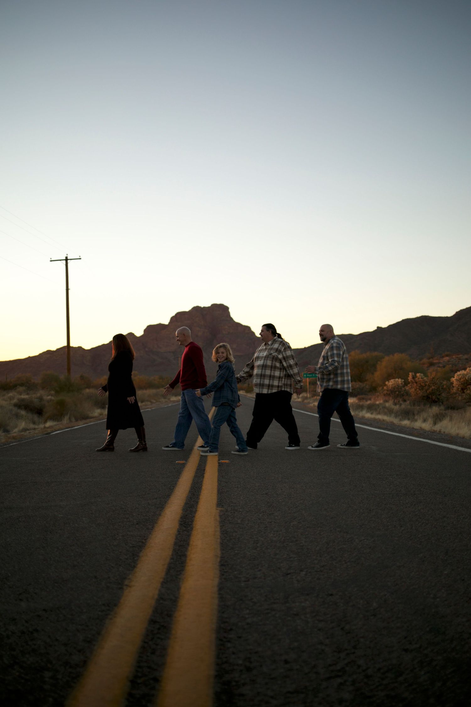 Five silhouettes walking across a desert road at sunset with mountains in the background.