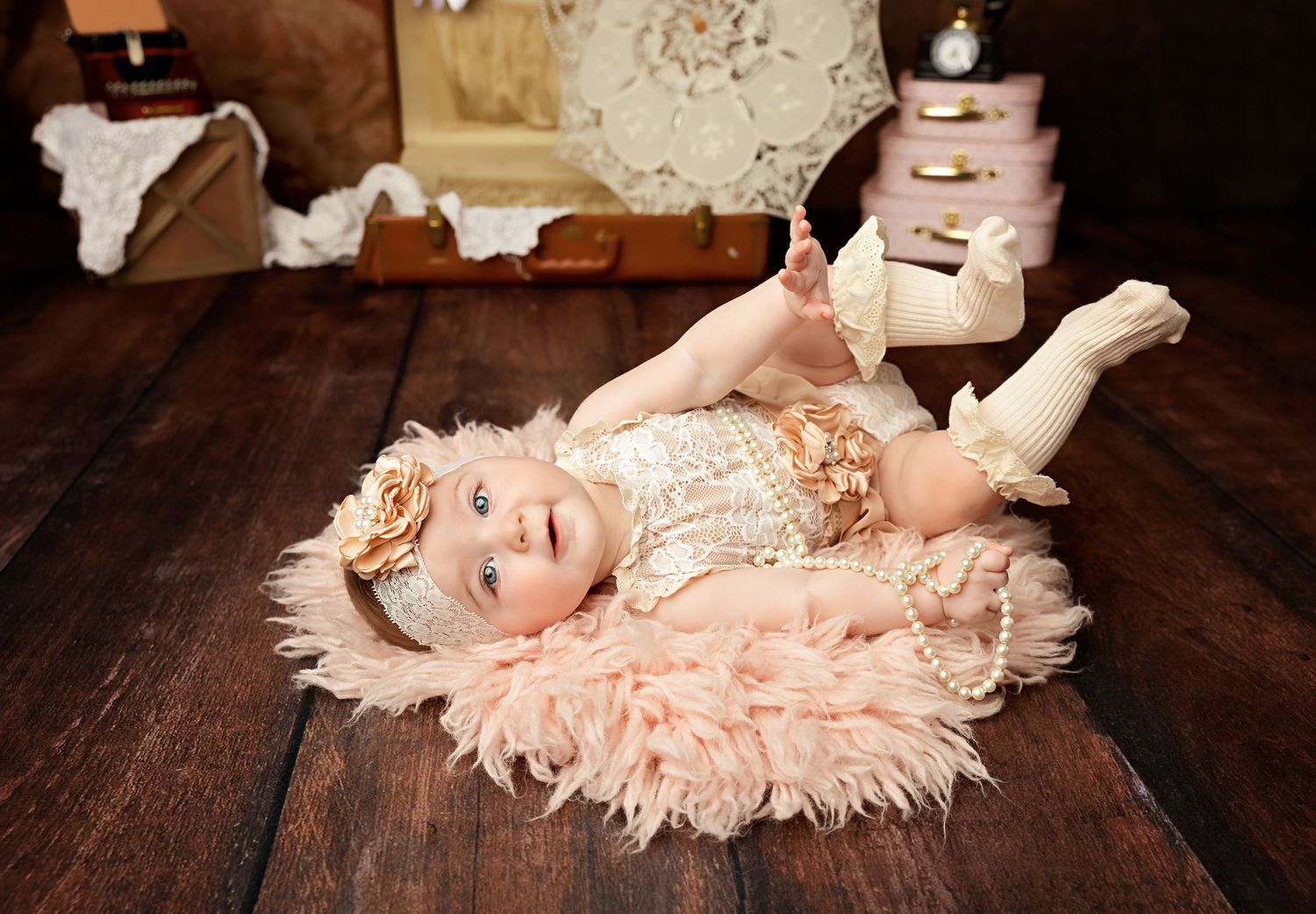Adorable baby girl in vintage lace romper lying on pink feather rug on rustic wood floor.