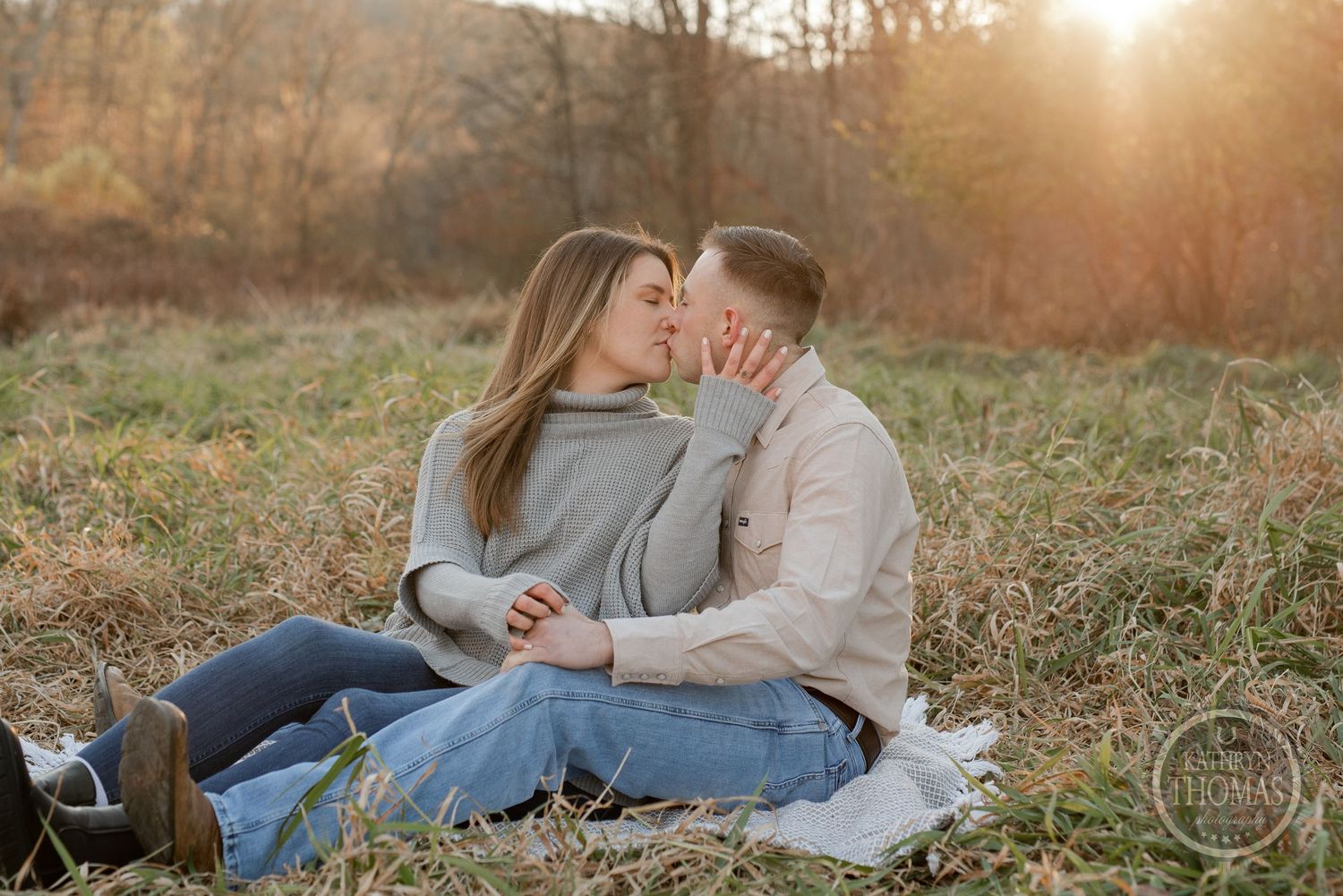 A couple sits on a blanket in a grassy field sharing an intimate moment at sunset.