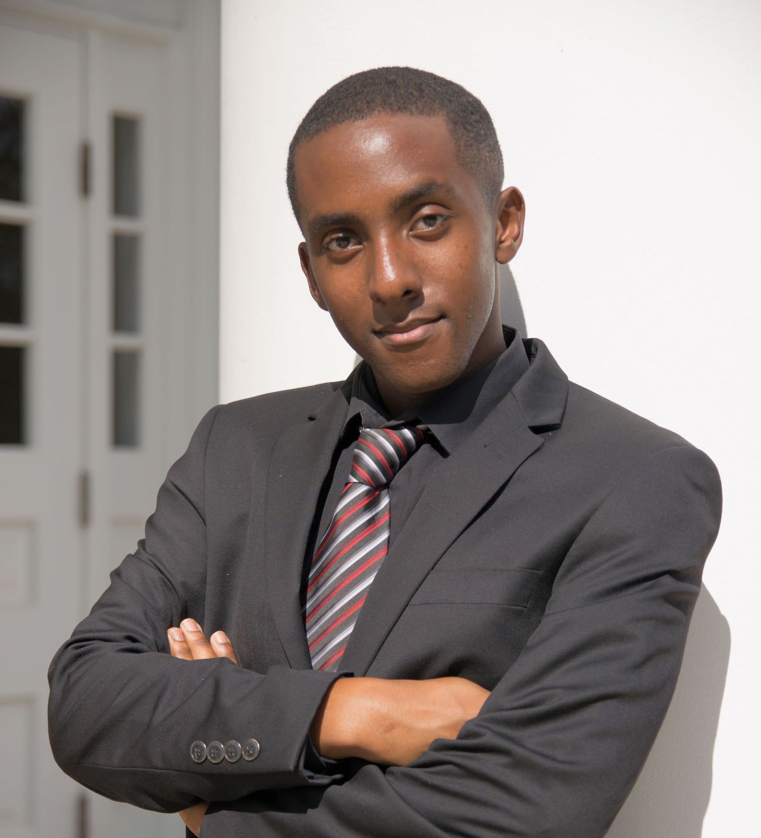 Professional in dark suit and striped tie poses with arms crossed against white background.