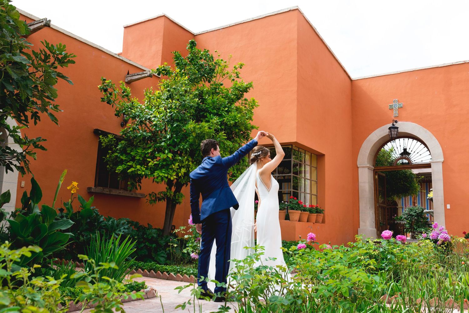 A couple poses in a garden courtyard with coral-colored walls and an arched entryway.