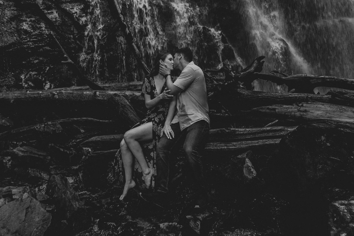 A couple sits intimately together near a waterfall in dramatic black and white photography.