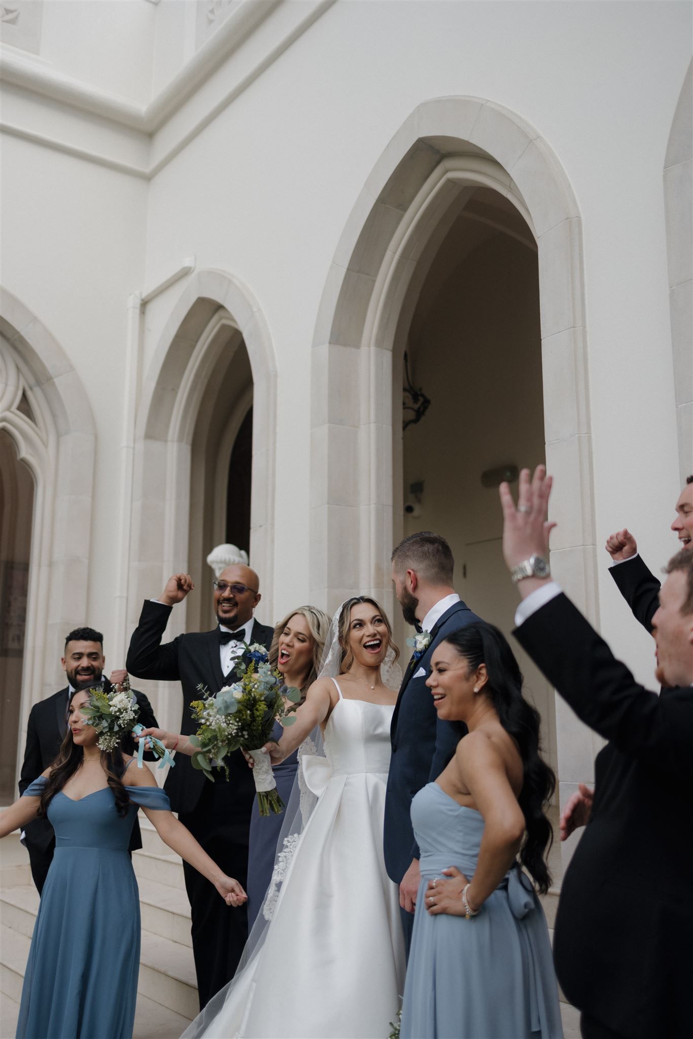 Wedding party celebrating beneath white arched entryway with bridesmaids in blue dresses and raised arms in joyful moment.