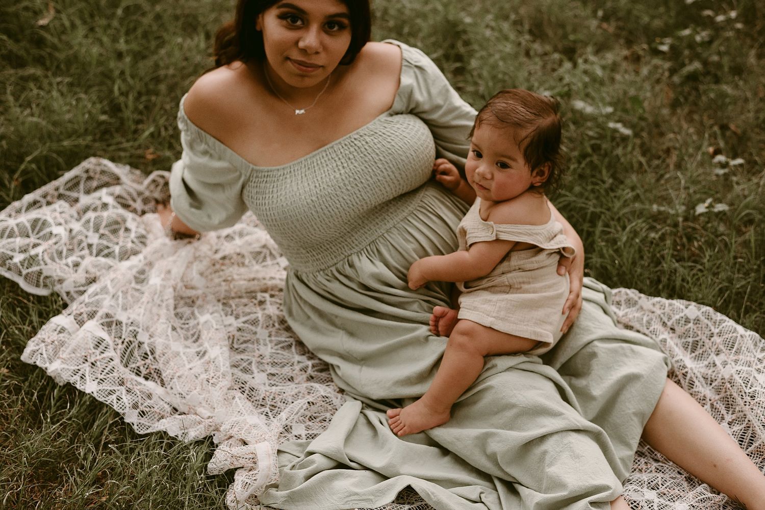 An intimate portrait of parent and child sitting together on white lace in a grassy outdoor setting.