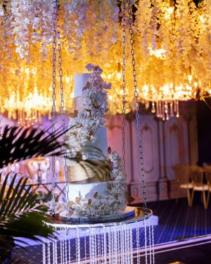 An ornate wedding cake displayed on a crystal stand under hanging crystal and floral decorations.
