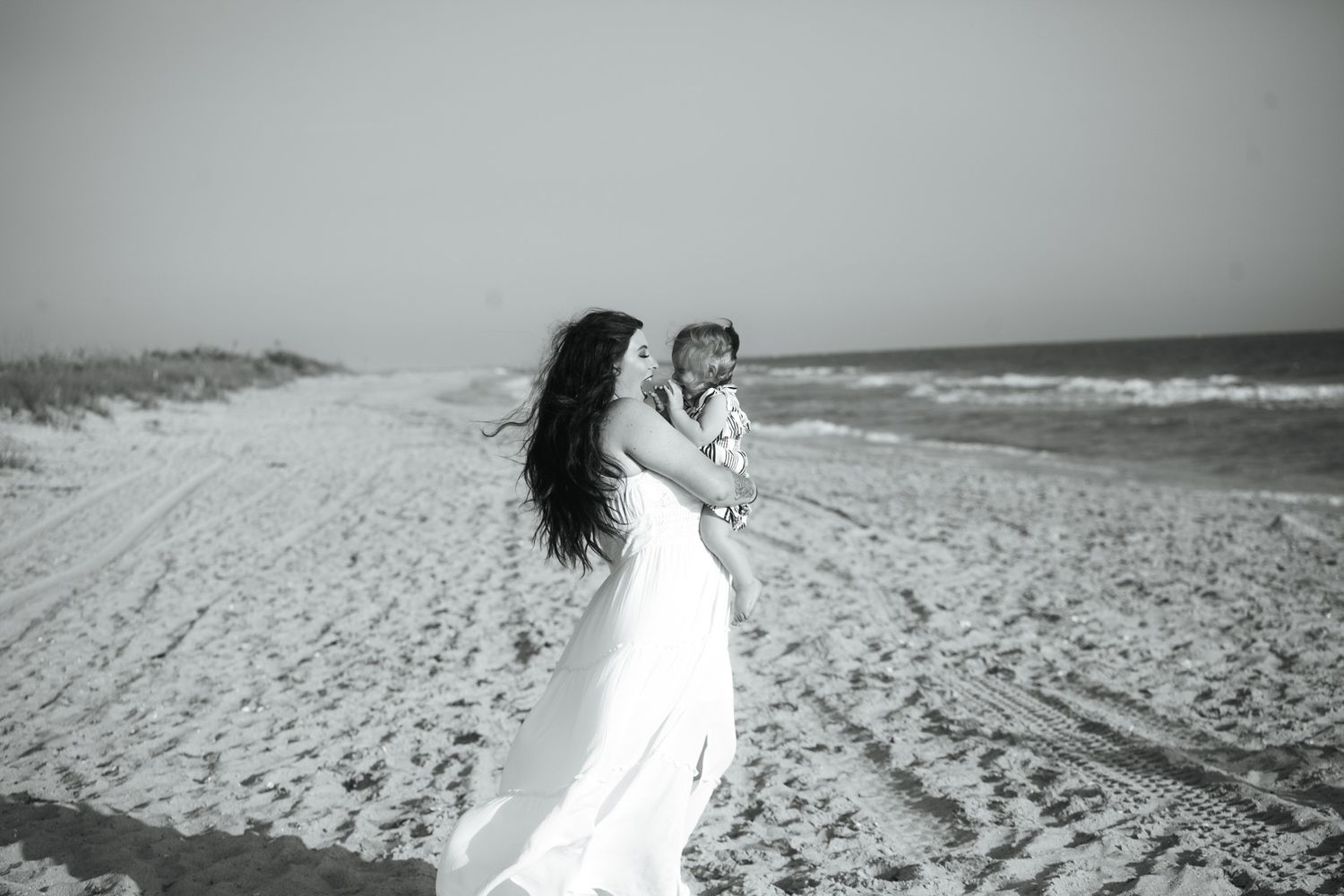 A woman in a white dress holding a baby on a sandy beach in black and white.