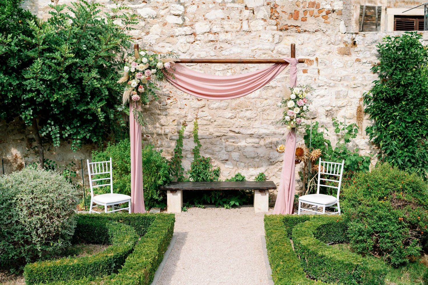 A rustic garden wedding arch with blush pink draping fabric and white chairs set against an old stone wall.