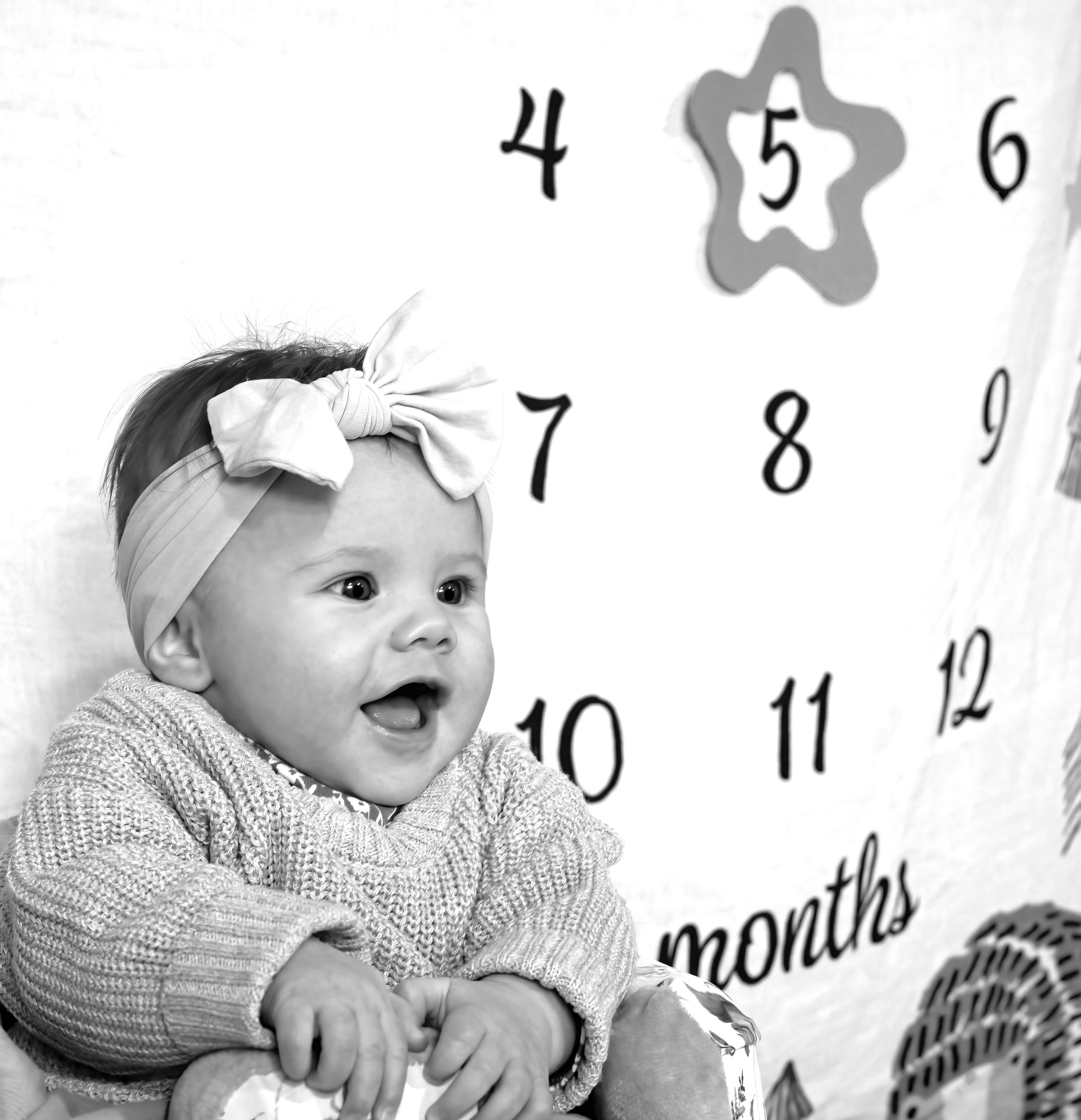 Baby in grey sweater with white bow headband smiling next to milestone month numbers.