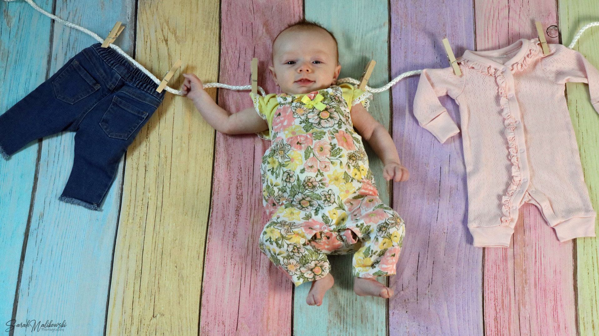 Baby growth milestone photos with pink and yellow clothes laid out on wooden floor from 1-12 months.