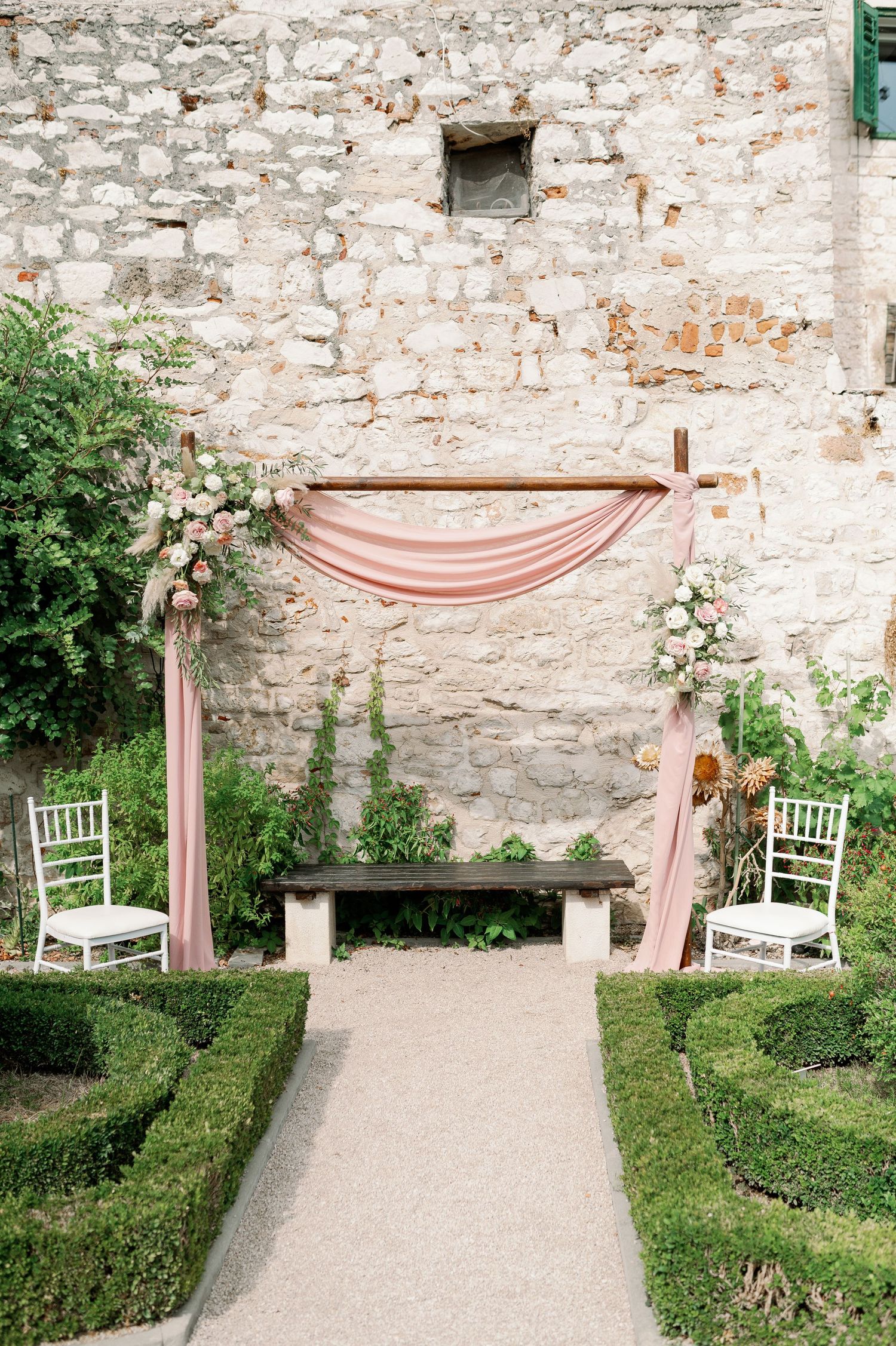 Garden wedding ceremony site with pink fabric arch and white chairs along manicured hedges.