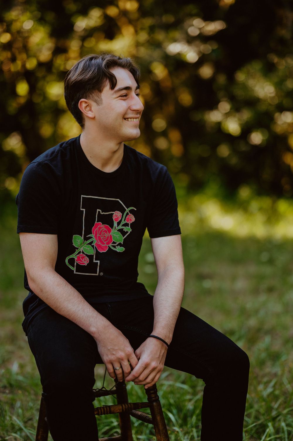 Person in black t-shirt smiles while sitting on wooden chair in natural outdoor setting.