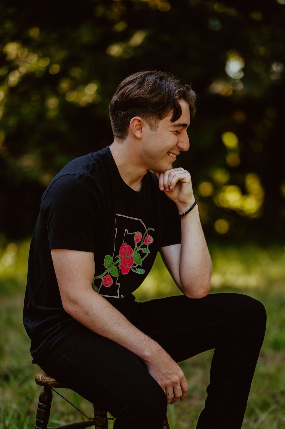 A person in black attire poses thoughtfully on a wooden stool against a sunlit natural background.