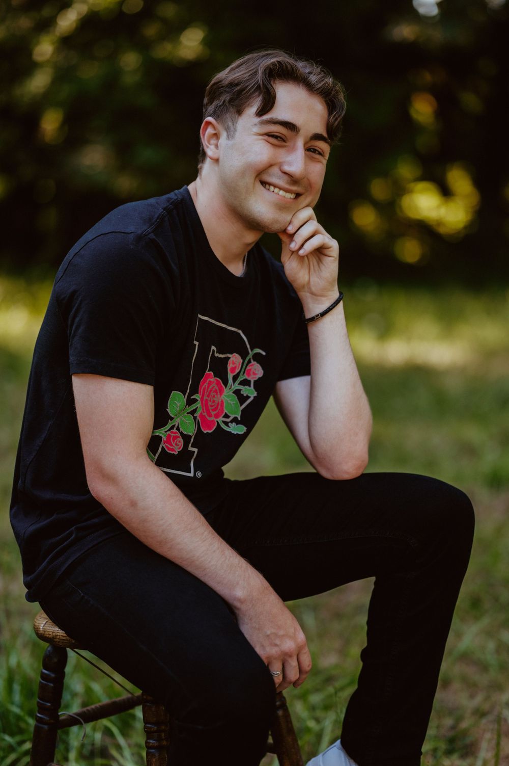A relaxed outdoor portrait of someone in black clothing sitting on a wooden stool with natural lighting.