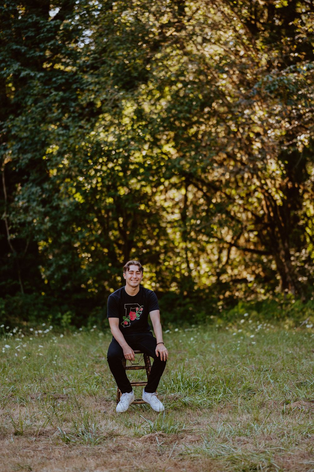 A full-length portrait of someone in black attire posed on a wooden stool in a sunny forest clearing.