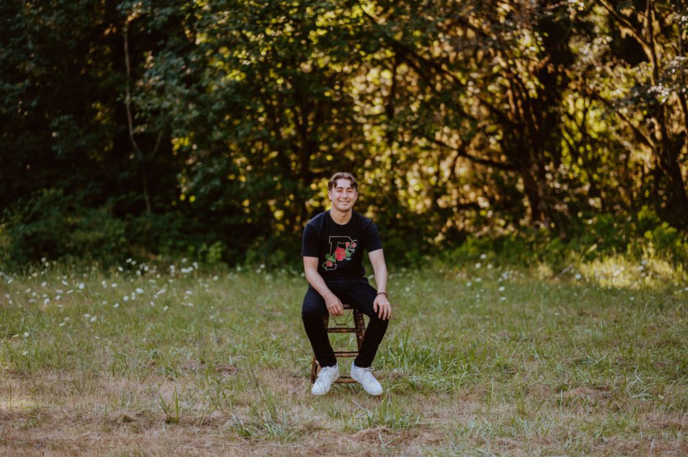 A person in dark clothing sits on a wooden stool in a sunlit meadow with trees in the background.