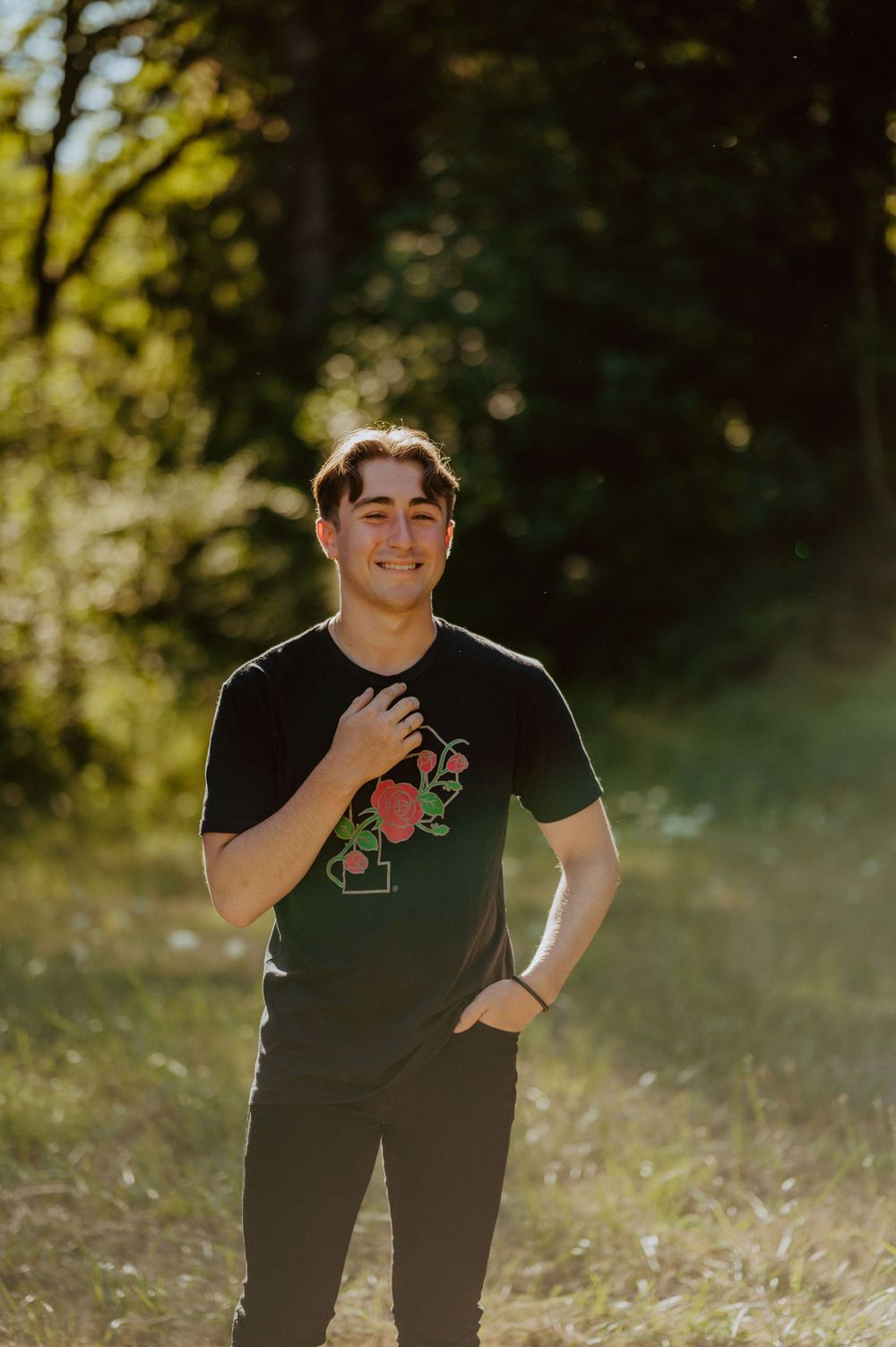 Someone poses in a black t-shirt with floral design against a sun-dappled forest background.