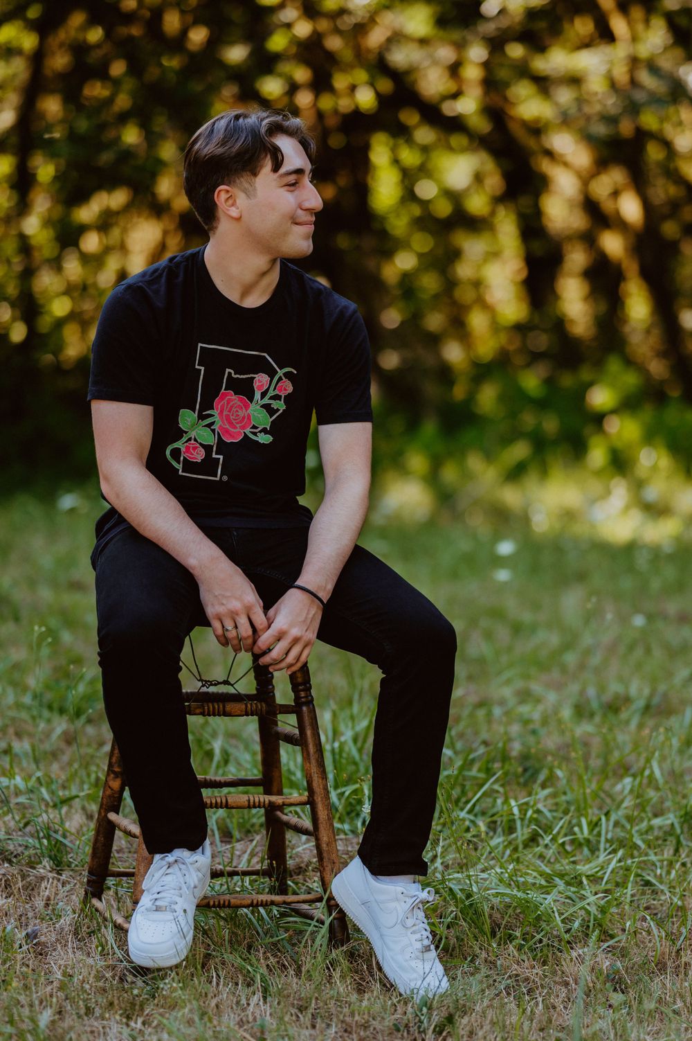 Person wearing a black t-shirt with rose design sits on wooden chair in sunny outdoor setting.