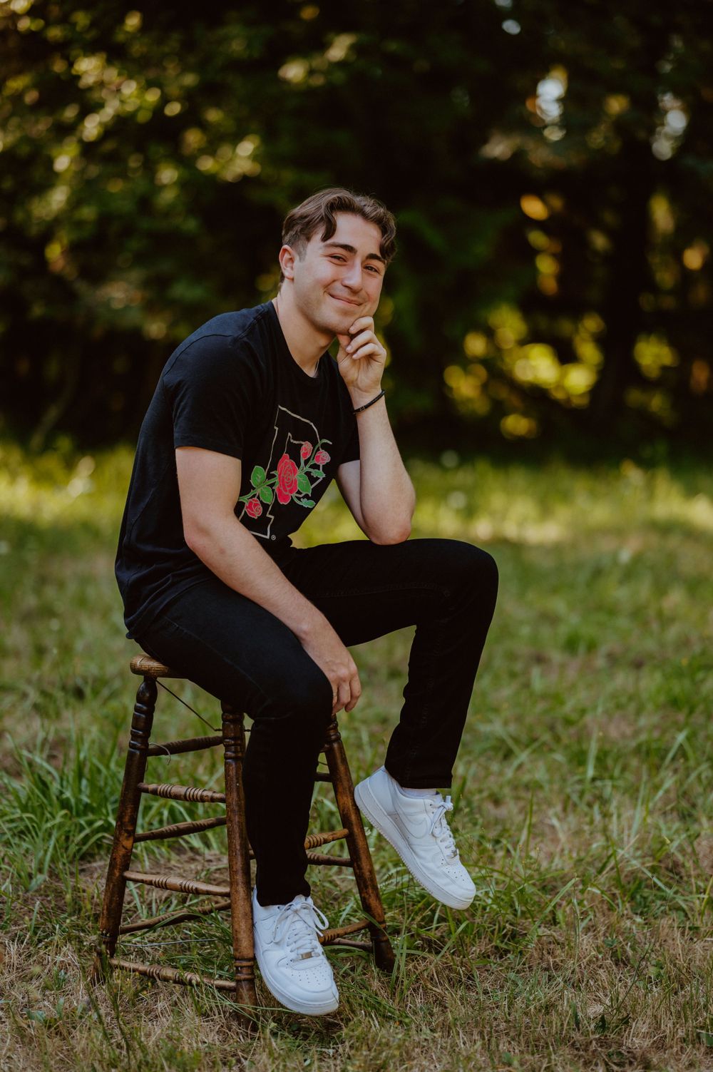 Someone sits casually on a wooden stool outdoors wearing a black t-shirt with floral design and white sneakers.
