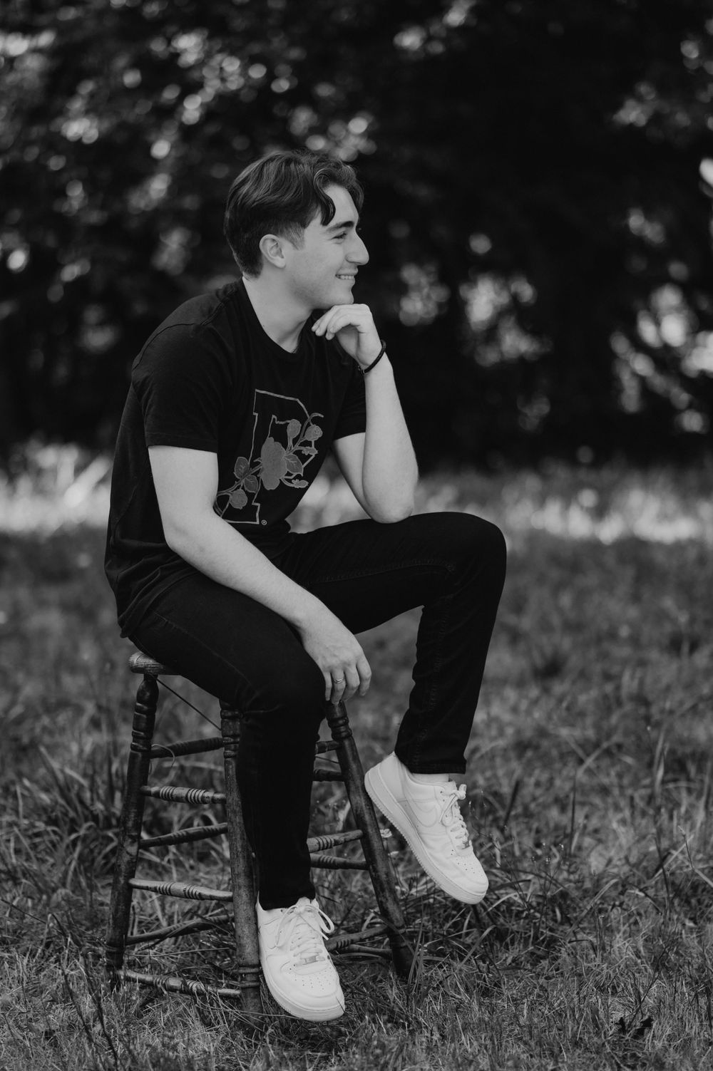 Black and white portrait of someone in casual attire sitting thoughtfully on a wooden stool outdoors.