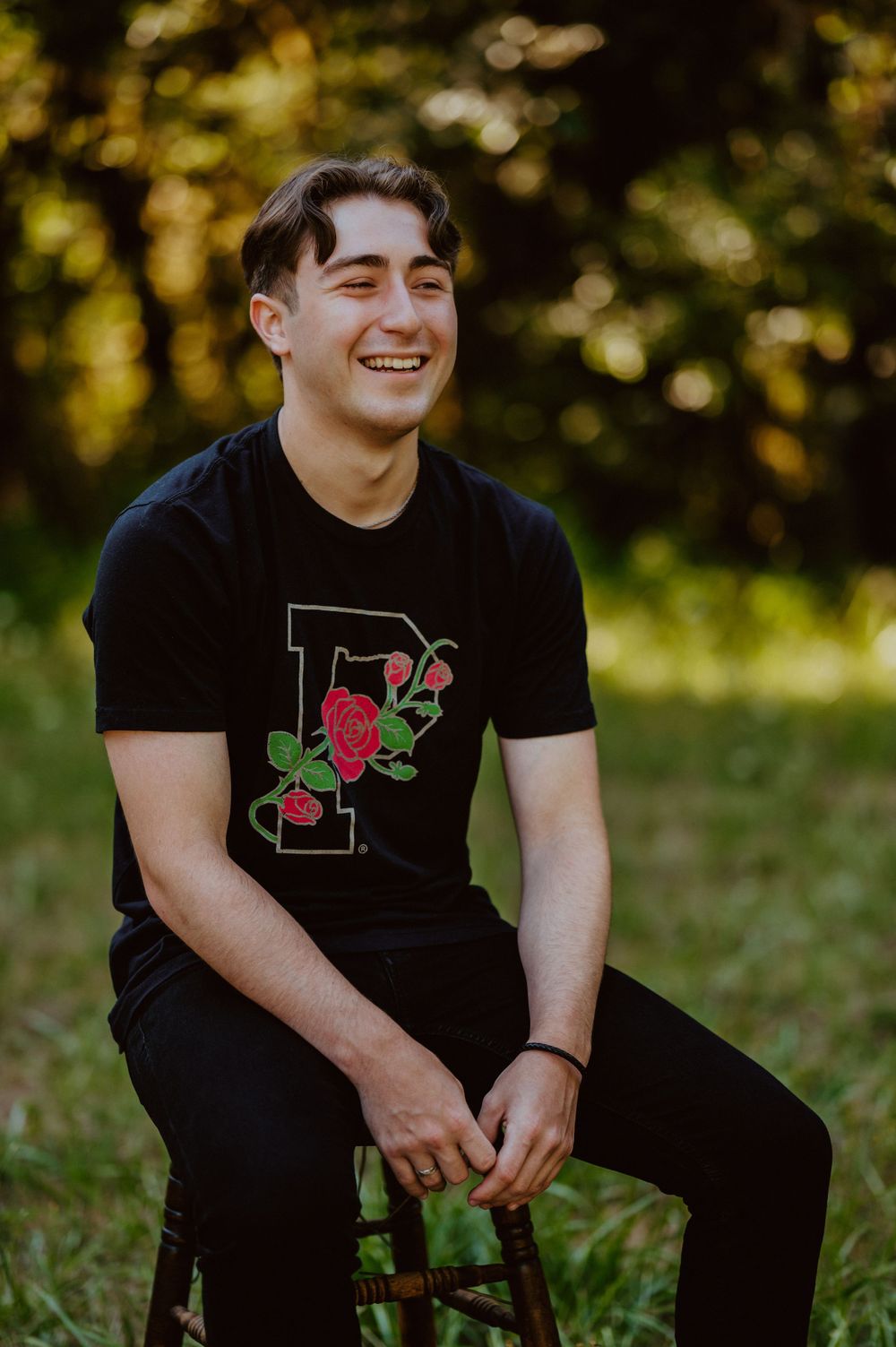 A person in a black t-shirt with a floral design sits on a wooden stool outdoors with a warm natural backdrop.