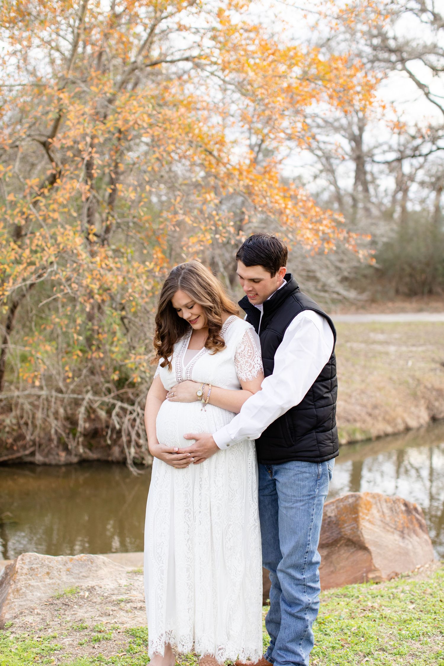 Couple embraces during fall maternity photo session near water.