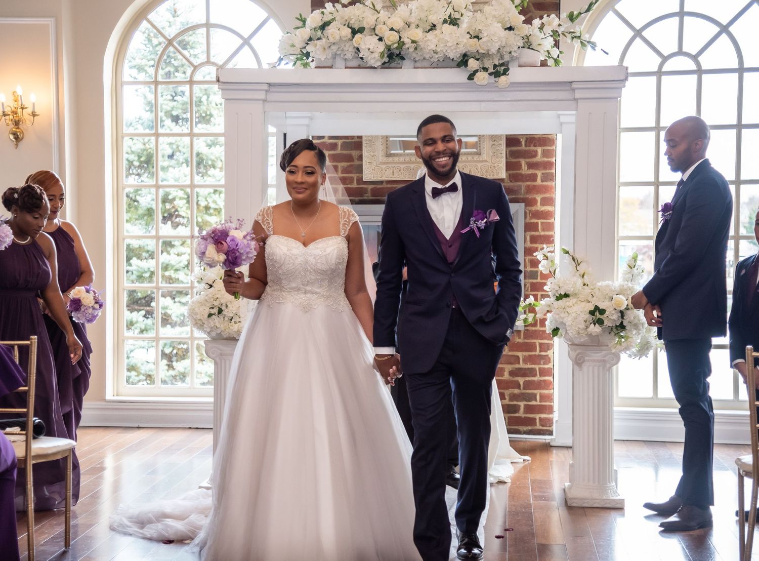 Elegant wedding ceremony in bright venue with floral arch and brick fireplace.