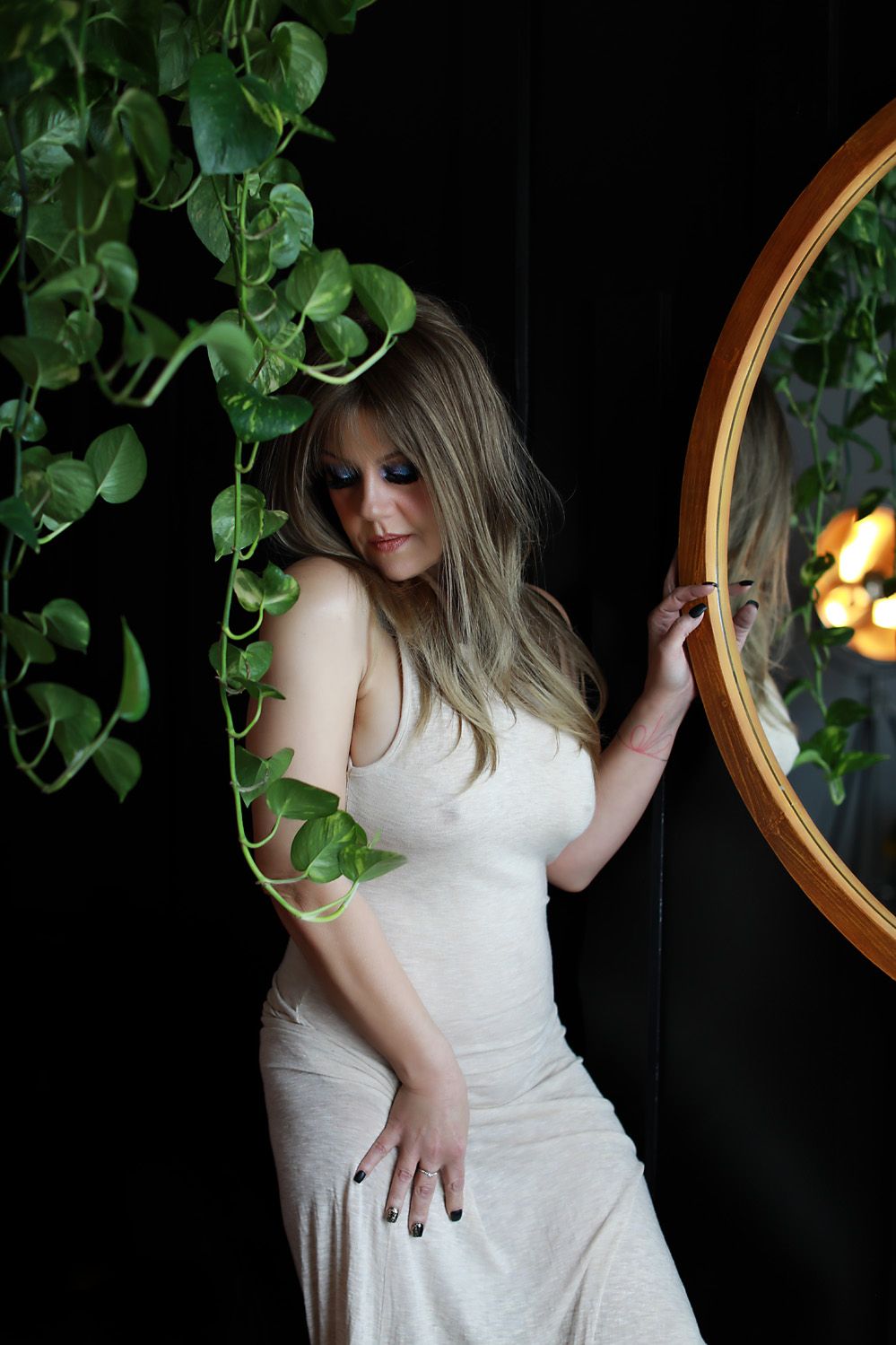Moody portrait with flowing white dress against dark wall with hanging plants and circular mirror in warm lighting.