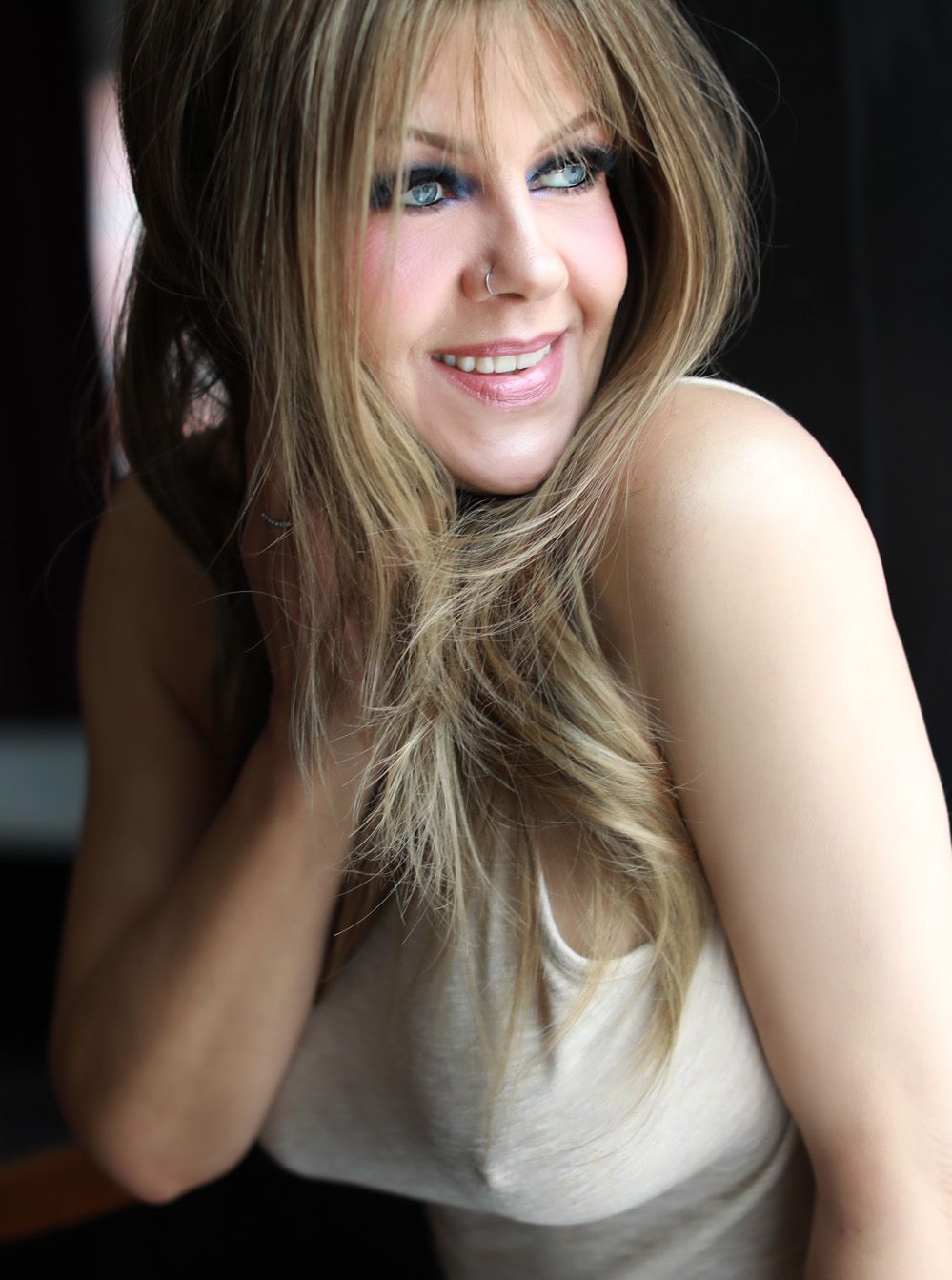 Portrait showing a natural smile, blonde hair swept to the side, and bright blue eyes against a dark background.
