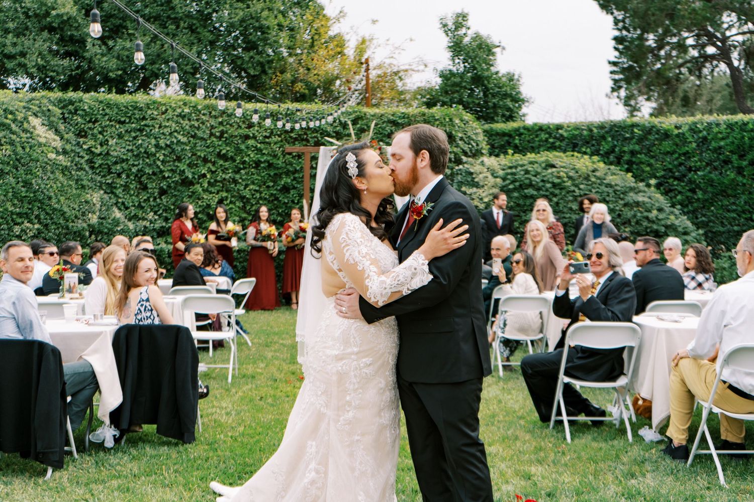 Newlyweds share first dance at outdoor garden reception with guests seated at tables.