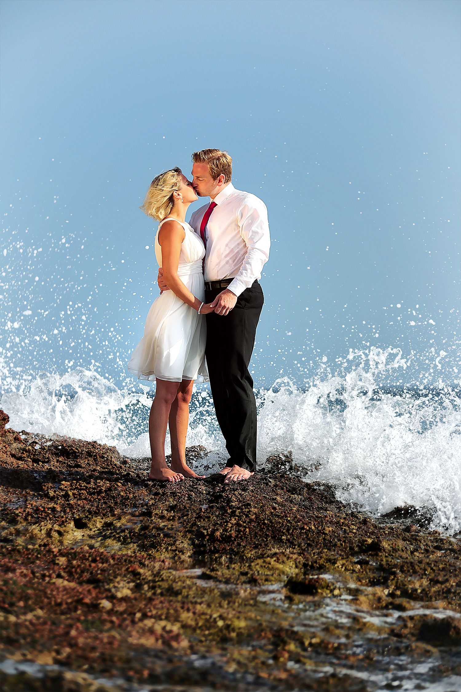 Couple kissing while ocean wave breakes at hey feet creating a visual spectacle in the air.