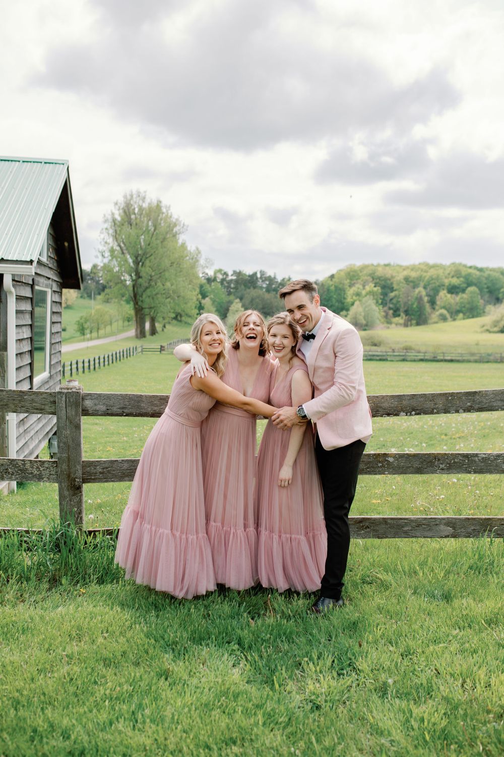 Wedding party in pink dresses and dark suit posing playfully by rustic wooden fence in countryside.