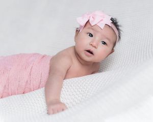 Baby in pink bow headband resting on soft white blanket.