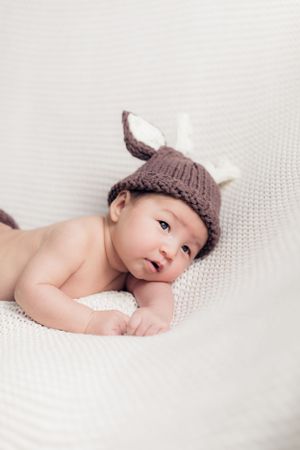 Adorable baby wearing brown knitted bunny hat lying on white textured fabric.