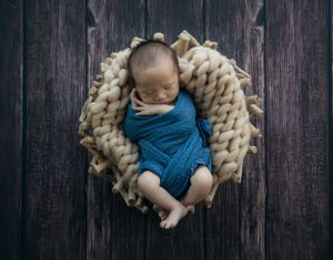 Sleeping newborn wrapped in blue swaddle on chunky knit basket.