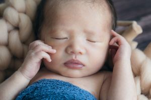 Sleeping newborn wrapped in blue blanket resting peacefully on cream-colored textured background.