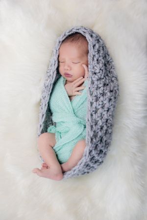 Sleeping newborn in mint green wrap nestled in gray knit cocoon on fluffy white background.