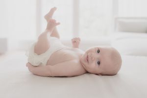 Baby wearing a diaper lying on back on white bedding while holding a toy.