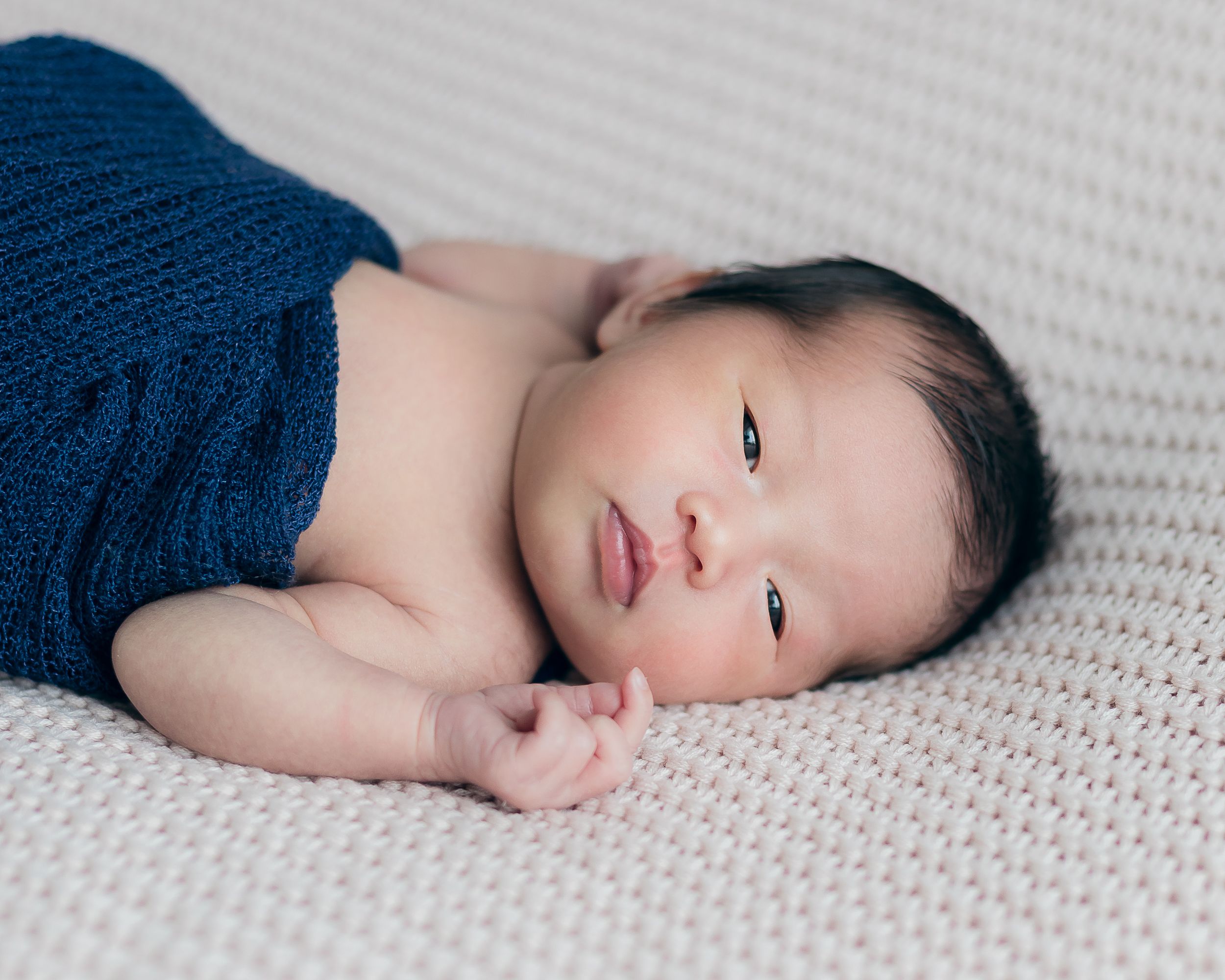 Baby wearing navy wrap lying on textured cream blanket.