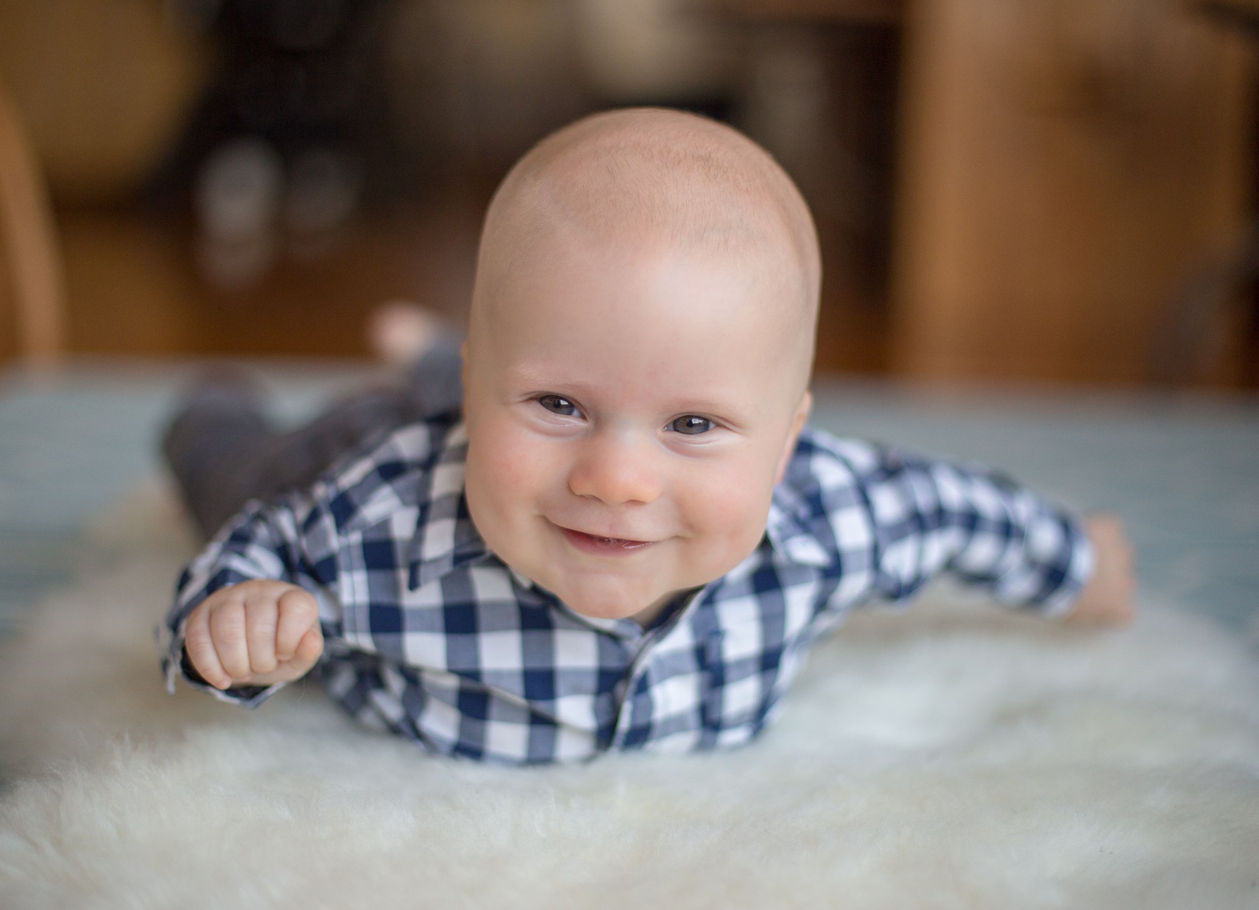 Baby in blue checkered shirt smiling while lying on fluffy white rug.