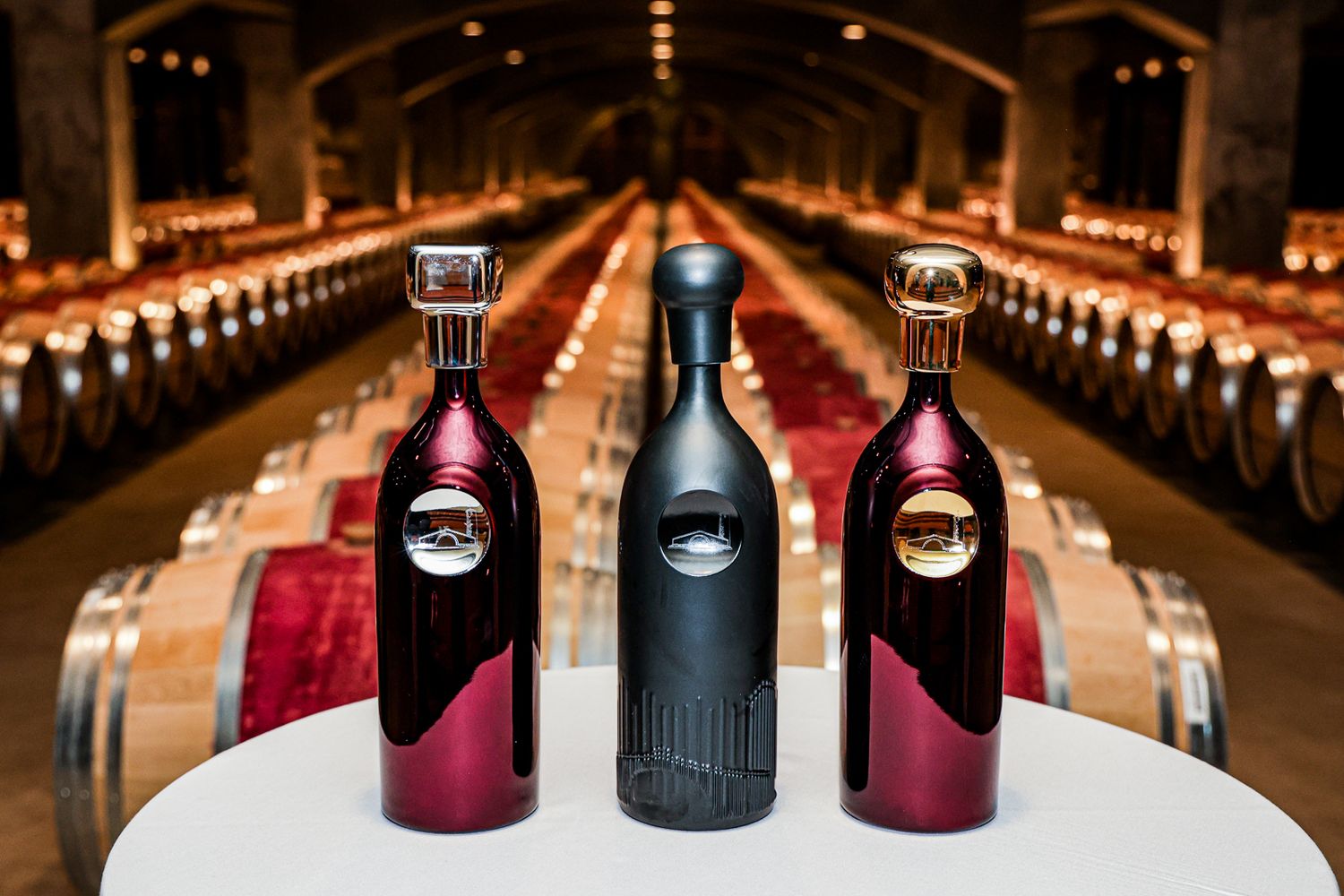 Three wine bottles displayed on a white table in a wine cellar with rows of barrels illuminated in the background.