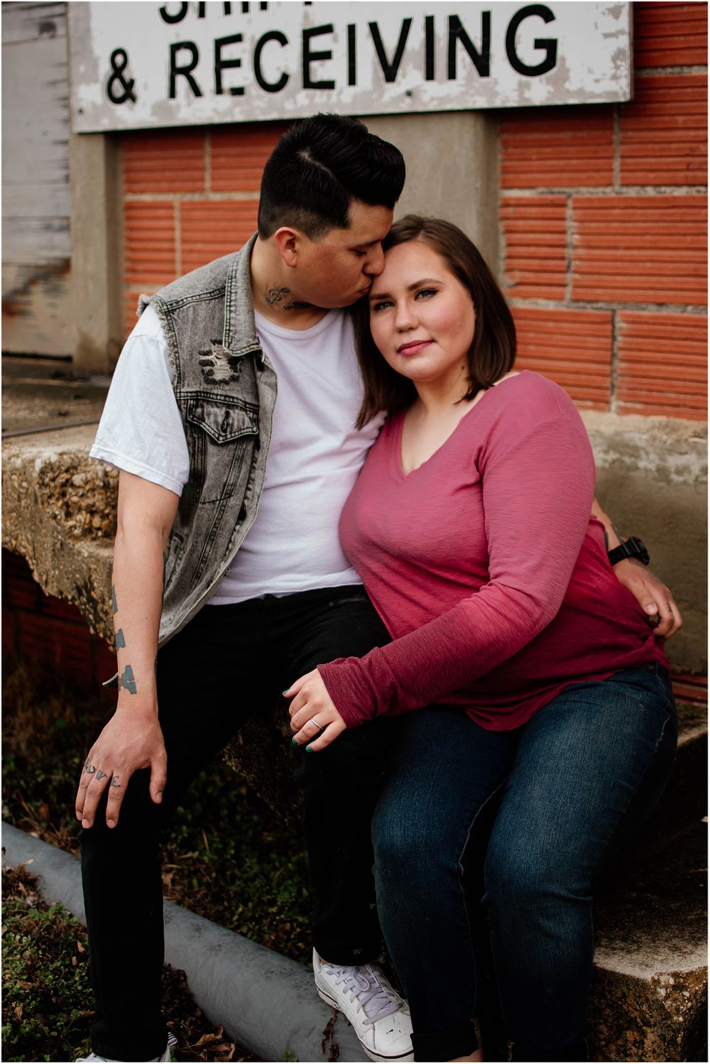 A couple poses together near a shipping and receiving sign on a brick wall.