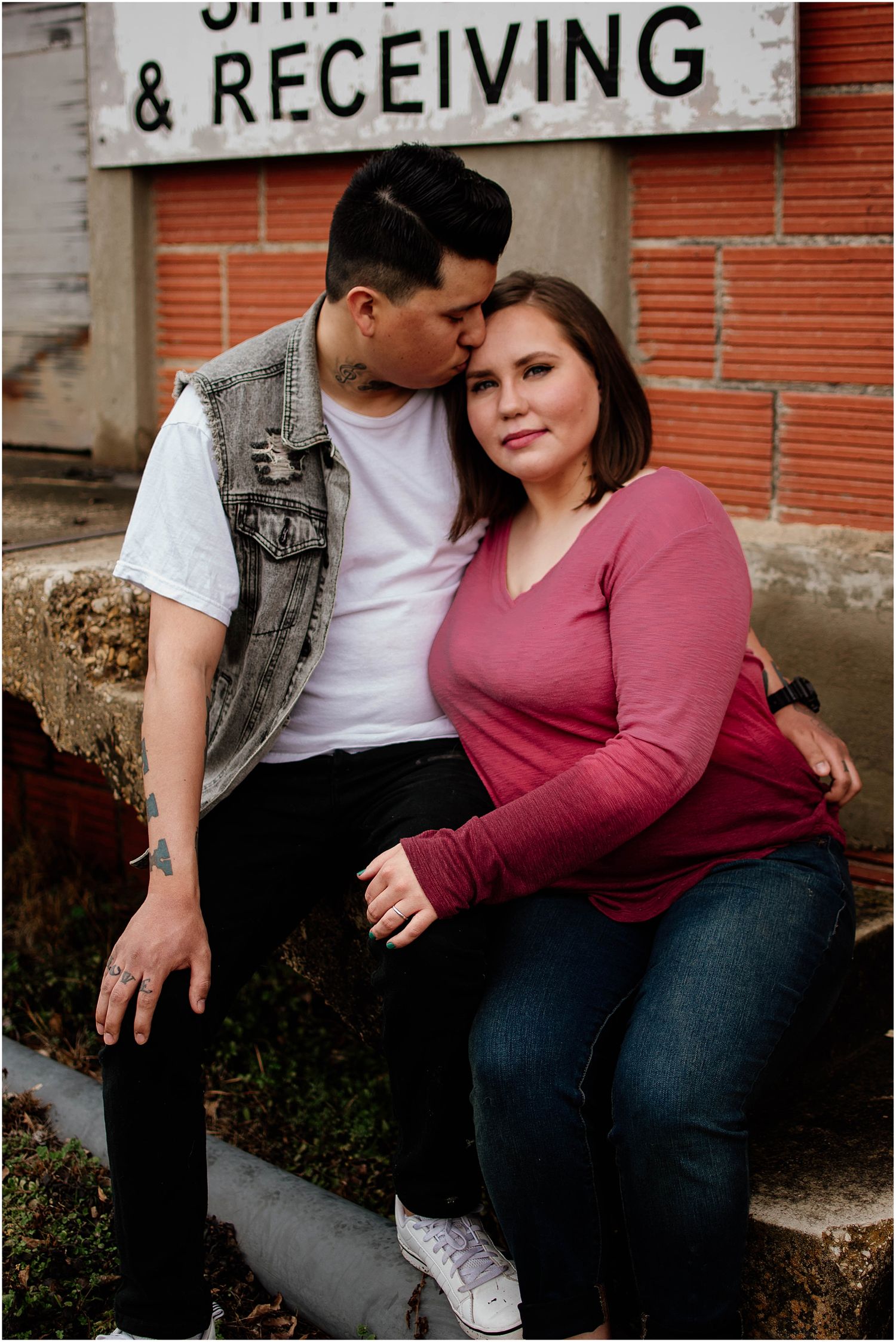 A couple poses together near a shipping and receiving sign on a brick wall.