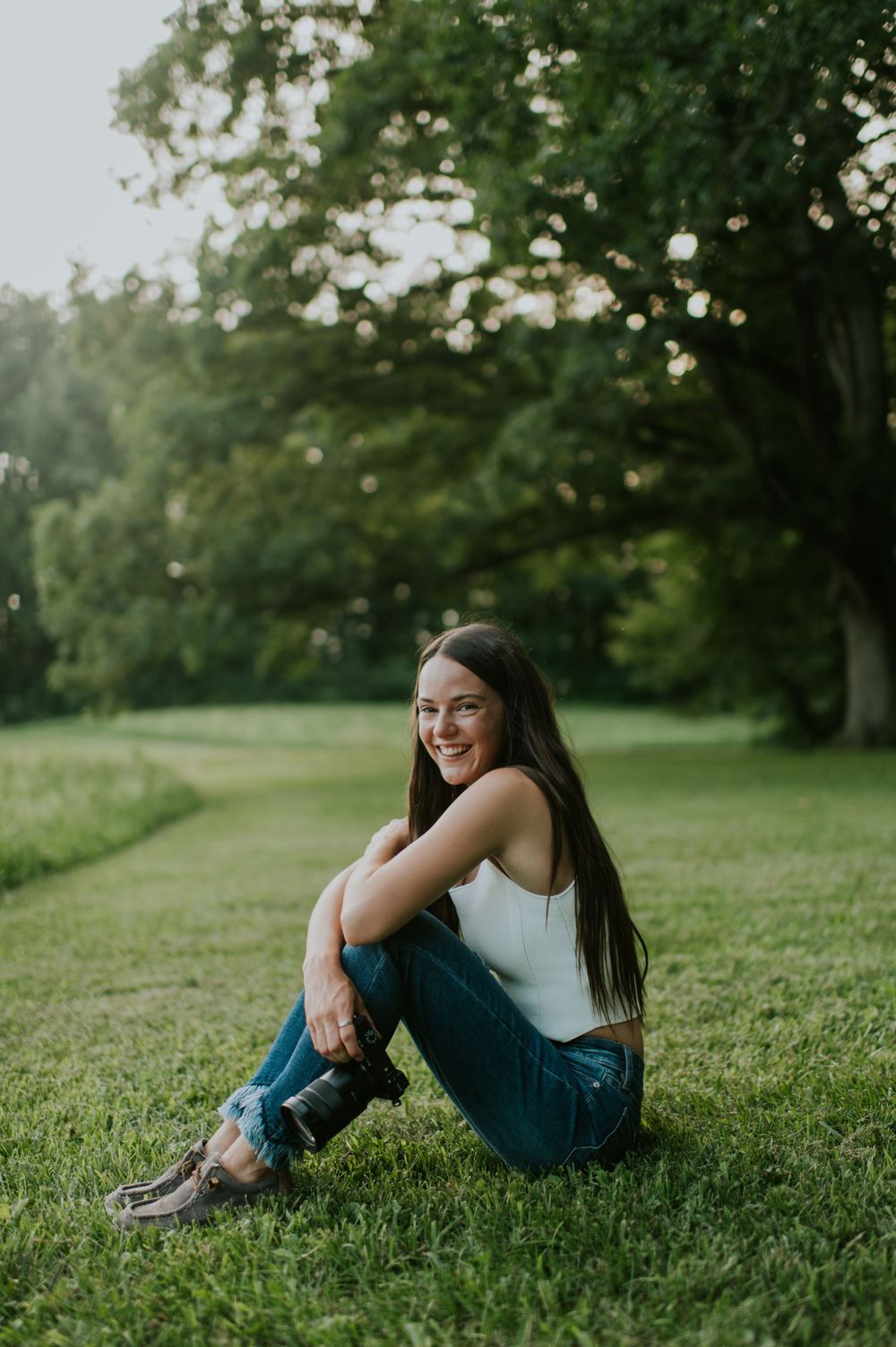 A person in white top and jeans sits on lush green grass with trees in the background.