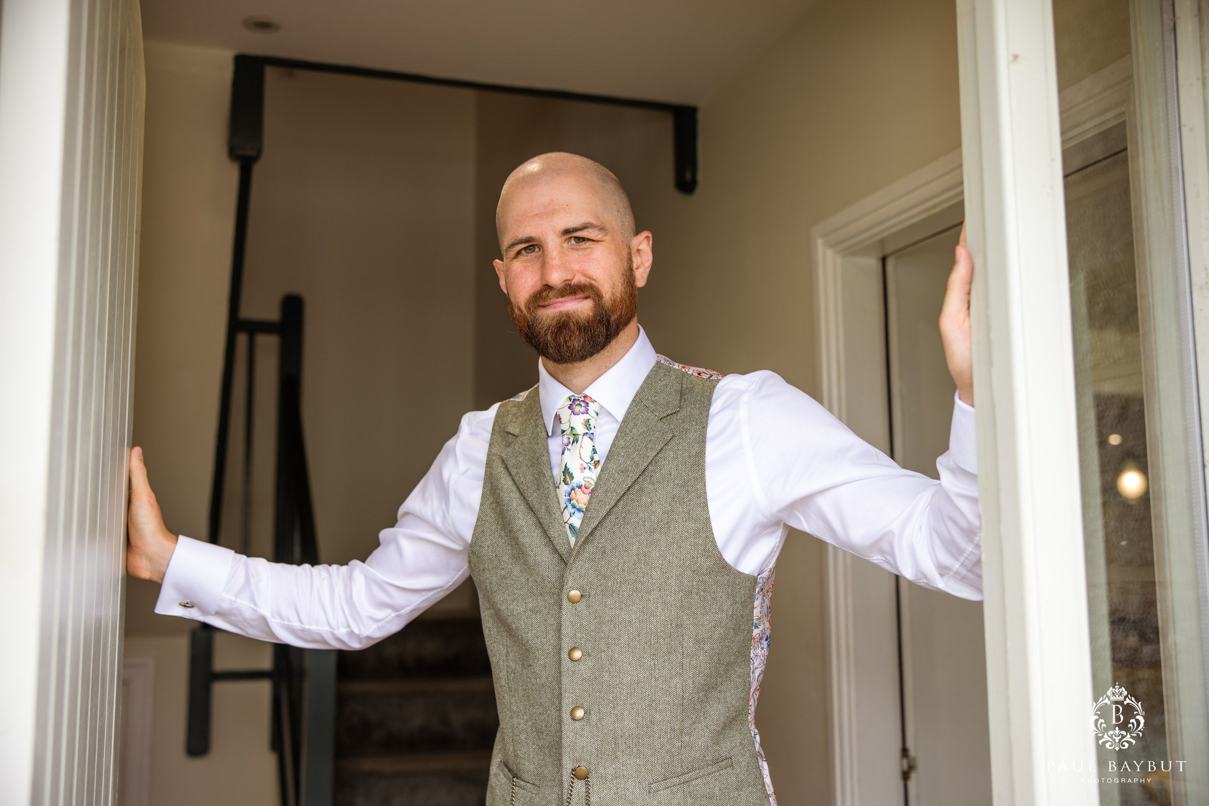 A groom in a grey vest and white shirt poses confidently in a doorway before his wedding ceremony.