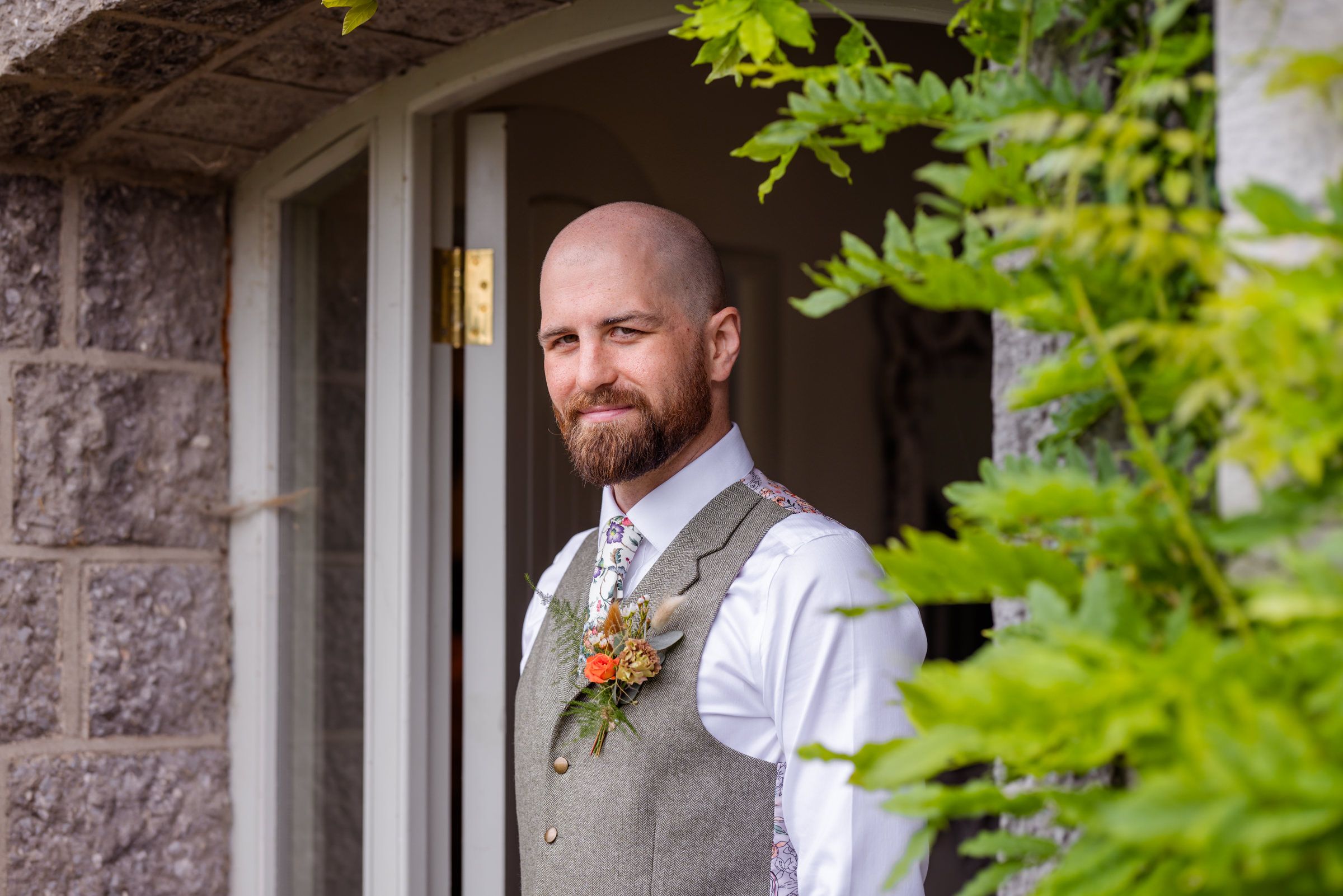 A wedding portrait showing a groom in a grey vest standing outside a stone building.